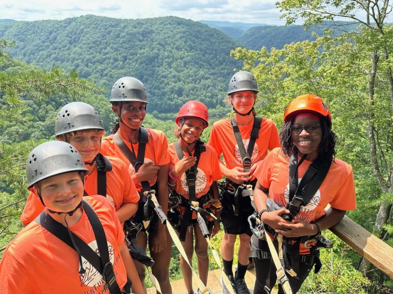 Five GEAR UP SWV students smiling with a GEAR UP SWV mentor smiling in front of green mountains while wearing a helmet and harness used for ziplining