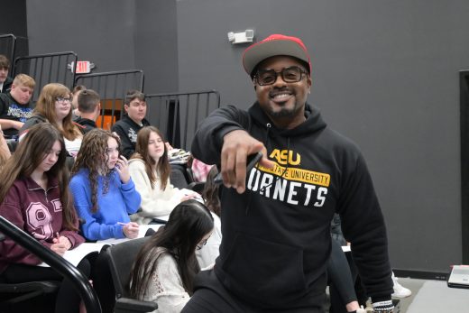 MJ Bridges points at the camera with a smile in front of students during a presentation about finances.