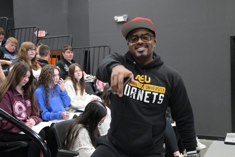 MJ Bridges points at the camera with a smile in front of students during a presentation about finances.